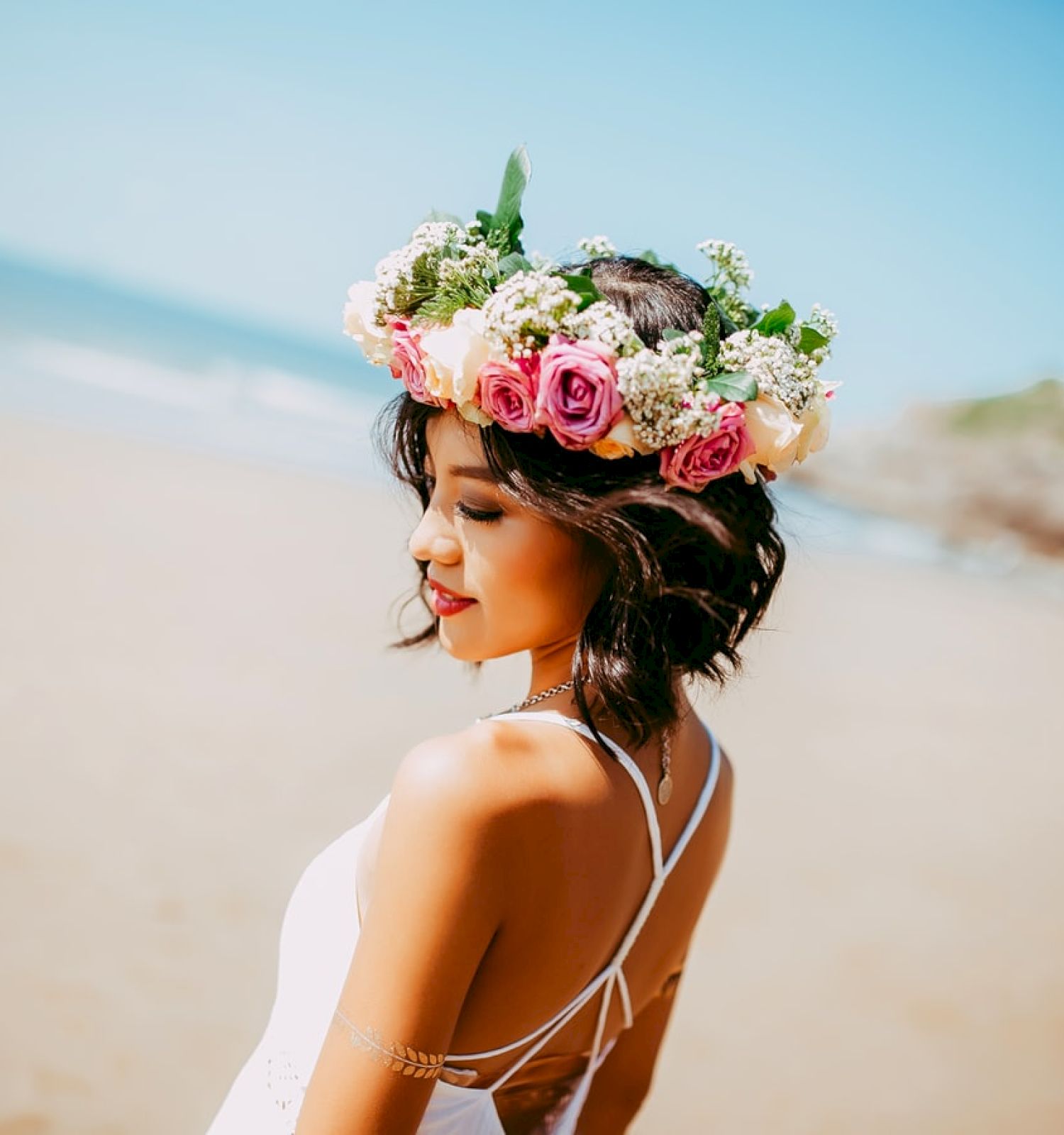 A woman wearing a floral crown and a white dress stands on a beach, with the ocean and blurred background in sight on a sunny day.