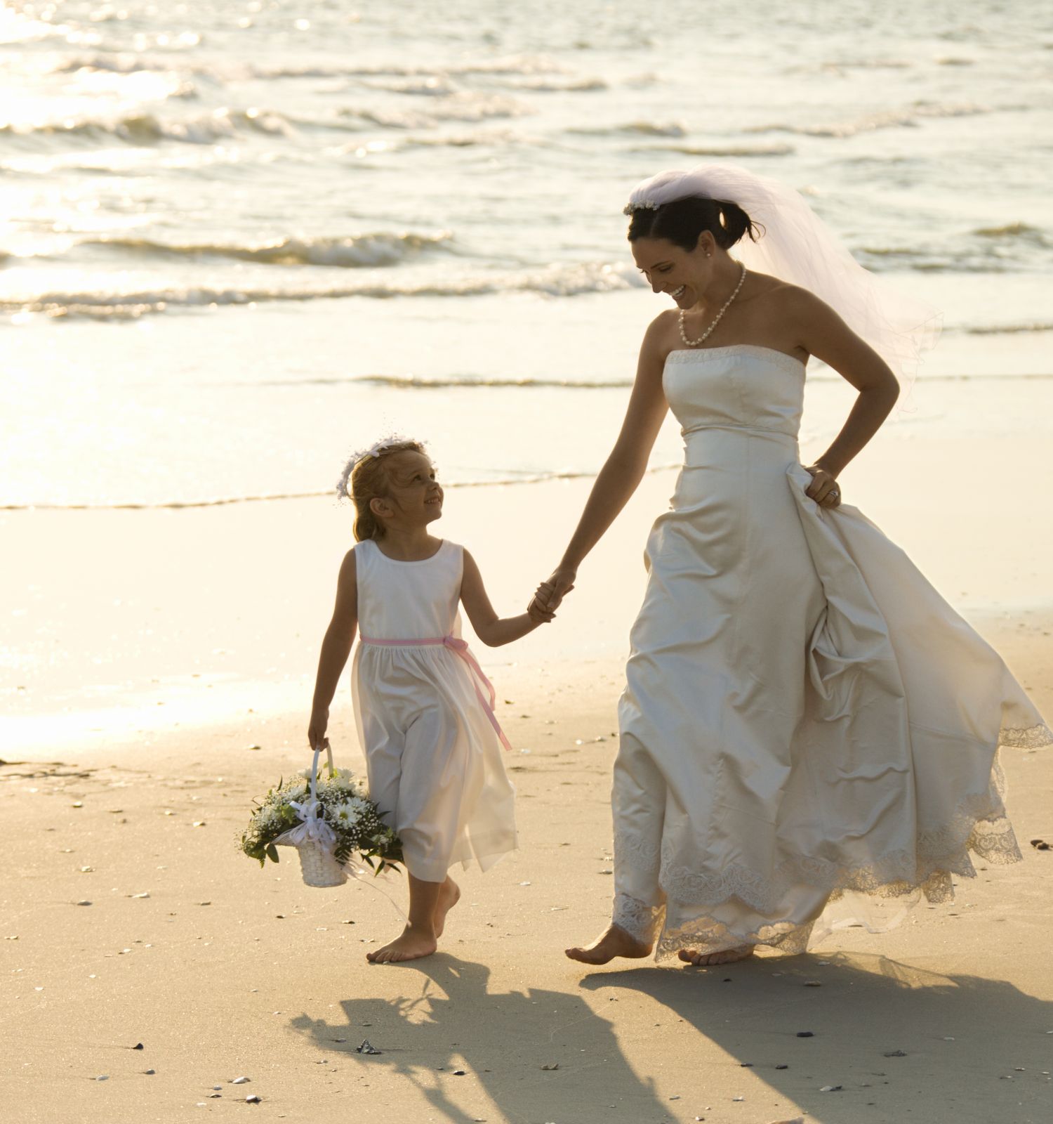 A bride and a young girl walk hand in hand on a beach; the girl holds a bouquet of flowers, and the sea is in the background.