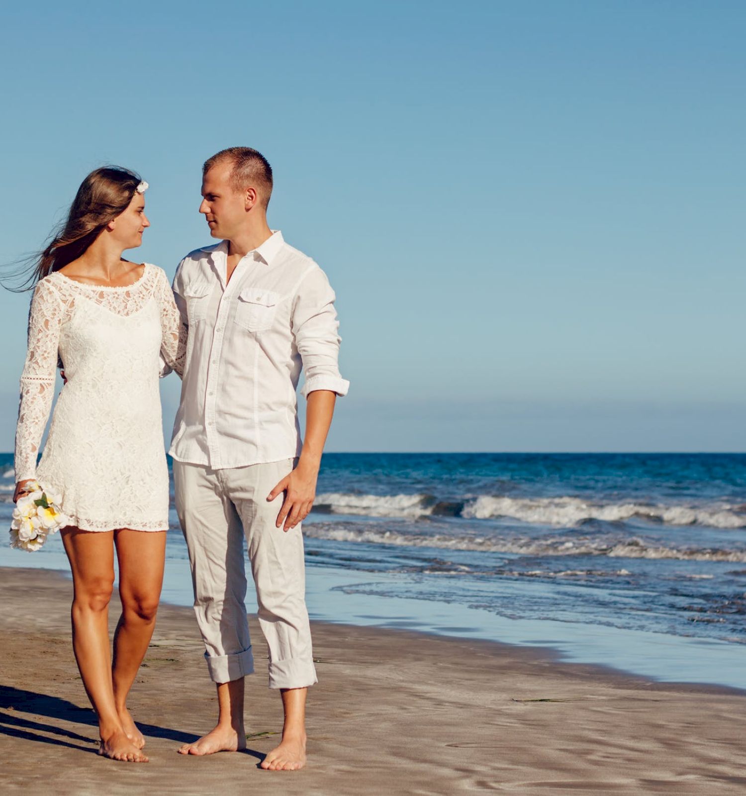 A couple is walking on the beach, holding hands and looking at each other. The woman wears a white dress and the man wears white clothes. The sky is clear.