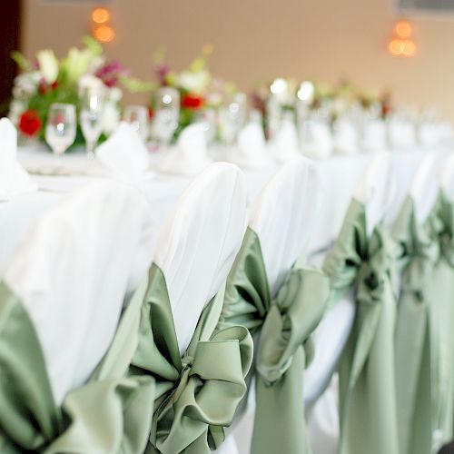 A decorated banquet table with white tablecloths, green chair sashes, floral arrangements, and glassware set up for a formal event or celebration.