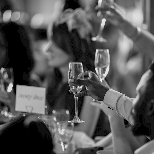 People are raising their glasses in a toast at a formal gathering, with a lit candle and table settings visible.