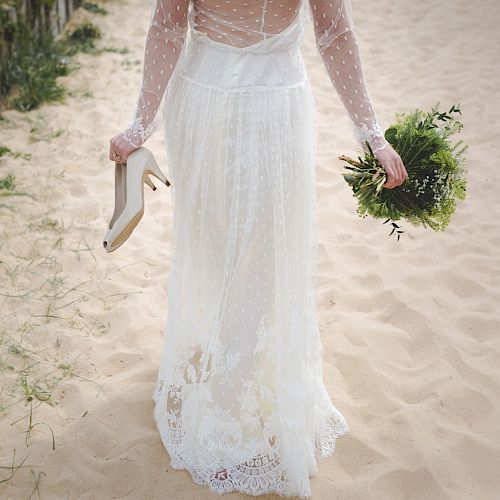 A person in a white wedding dress walks on the sand, holding their shoes in one hand and a bouquet in the other, near a grassy area.