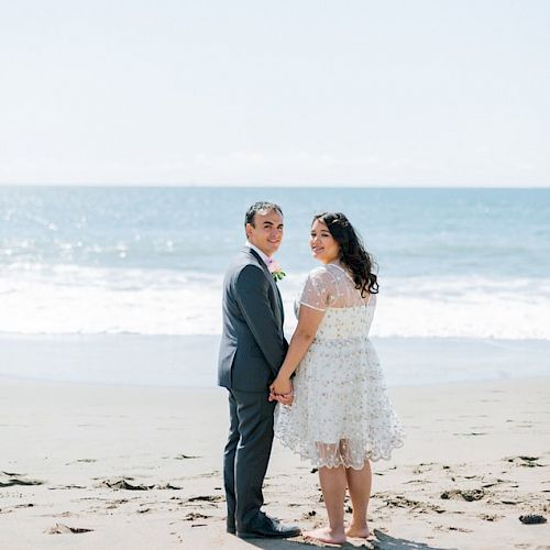 A couple is standing on a beach, holding hands and facing the camera, with the ocean and a clear blue sky in the background.