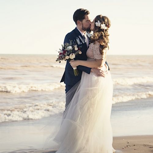 A couple in wedding attire shares a kiss on a beach, with the ocean waves and sandy shore in the background, capturing a romantic moment.