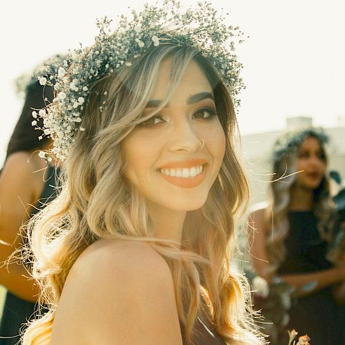 A woman with long, wavy hair and a floral crown smiles warmly. Other similarly adorned women are visible in the background, suggesting a joyful event.