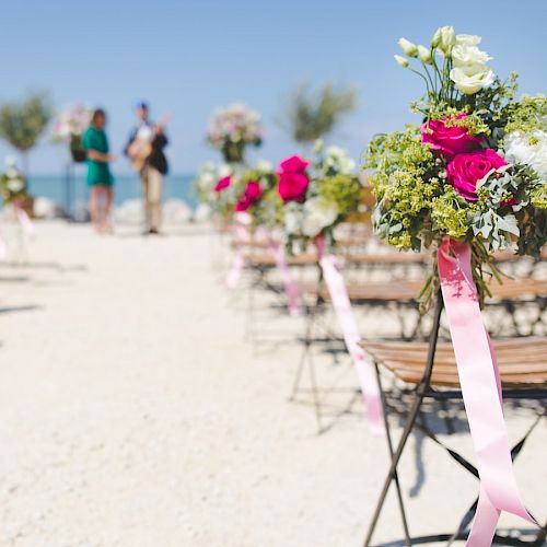 This image shows an outdoor wedding setup with rows of chairs adorned with flower bouquets, and blurred figures in the background near the altar.