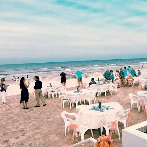 A beachside event with tables and chairs set up, people mingling, and a scenic ocean view in the background.