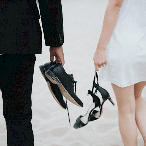 Two people, one in a black suit and the other in a white dress, walk on the beach carrying their shoes in hand.