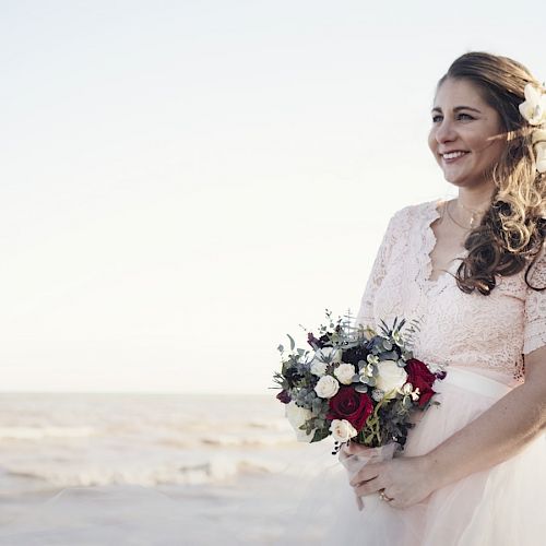 A smiling woman in a lace dress holds a bouquet, standing by the seaside with gentle waves in the background, and a flower in her hair.