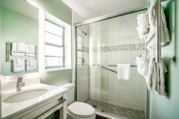 The image shows a modern bathroom with a white vanity, a toilet, and a glass-enclosed shower. Towels are neatly hung on a rack beside the shower.