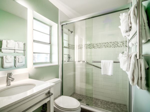The image shows a modern bathroom with a white vanity, a toilet, and a glass-enclosed shower. Towels are neatly hung on a rack beside the shower.