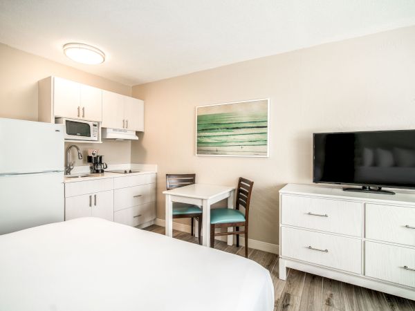 The image shows a clean, modern studio apartment with a kitchenette, dining area, bed, flat-screen TV on a dresser, and wall art.