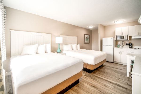 The image shows a hotel room with two queen beds, a bedside table, a lamp, a mini kitchen, and light wood flooring, designed in a modern style.