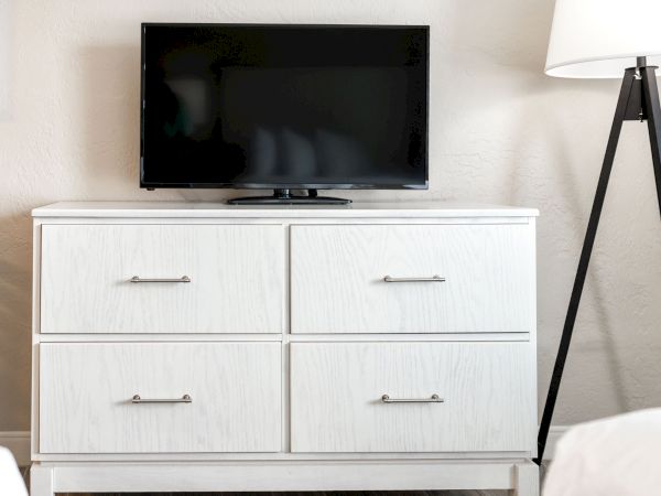 A small flat-screen TV is placed on top of a white dresser, which has four drawers, next to a floor lamp with a tripod base.