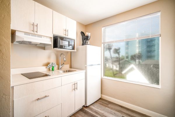 The image shows a bright, small kitchenette with a stove, sink, refrigerator, microwave, and coffee maker, in front of a window with blinds.