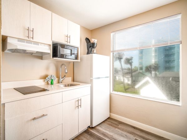The image shows a bright, small kitchenette with a stove, sink, refrigerator, microwave, and coffee maker, in front of a window with blinds.