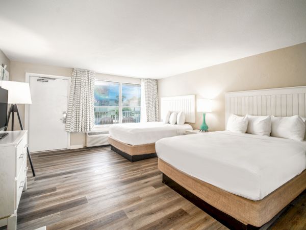 A clean, bright hotel room with two double beds, wooden flooring, a flat-screen TV, a window with curtains, and bedside tables with lamps.