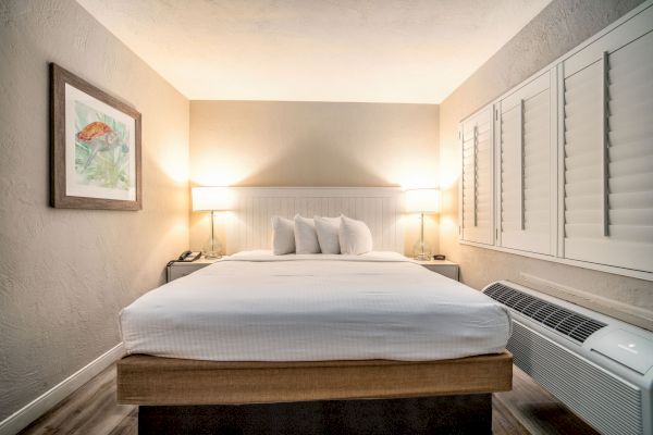 A well-lit hotel room with a neatly made bed, bedside lamps, wall art, and a window with shutters and an air conditioning unit below it.