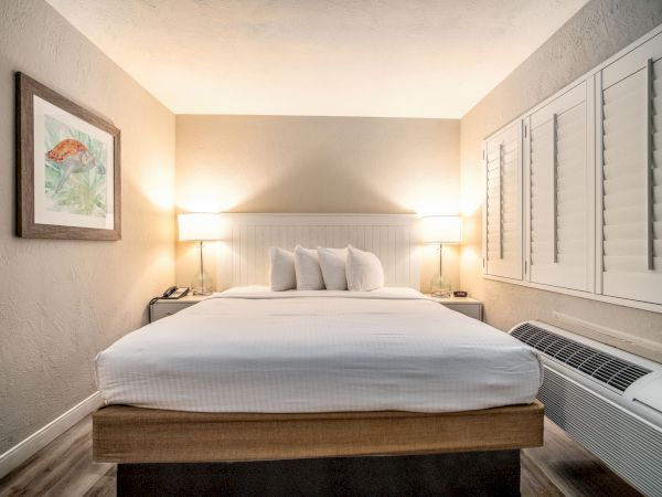 A well-lit hotel room with a neatly made bed, bedside lamps, wall art, and a window with shutters and an air conditioning unit below it.