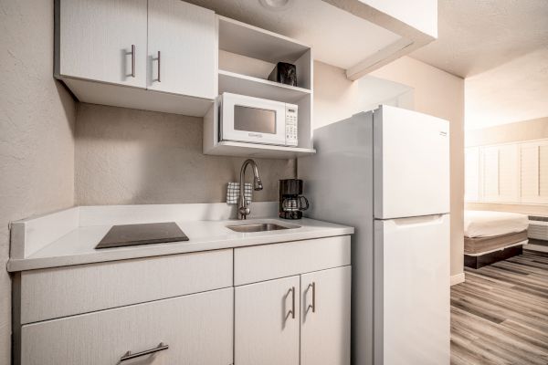 A compact kitchen with white cabinets, a microwave, stovetop, sink, coffee maker, and refrigerator is shown, adjacent to a bedroom area.