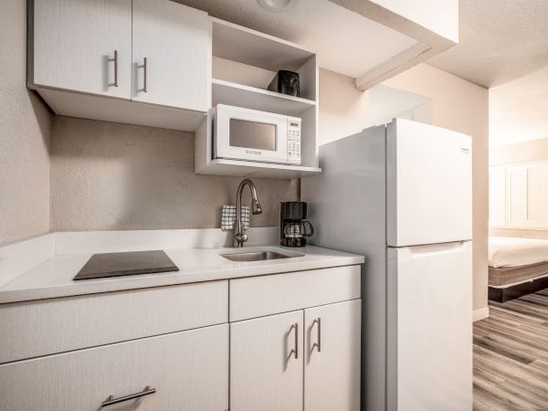 A compact kitchen with white cabinets, a microwave, stovetop, sink, coffee maker, and refrigerator is shown, adjacent to a bedroom area.