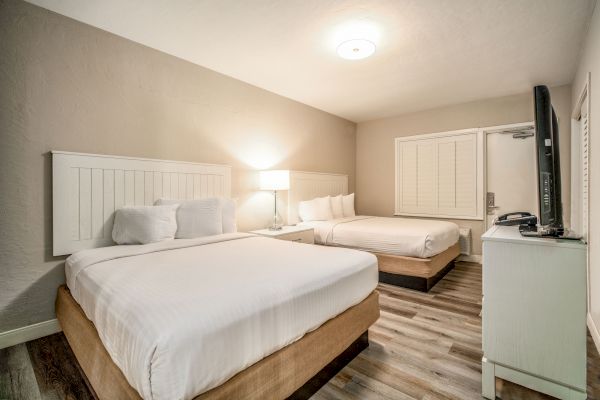 The image shows a neatly arranged hotel room with two double beds, bedside lamp, a flat-screen TV on a dresser, and wooden flooring.