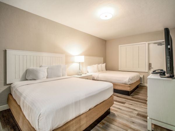 The image shows a neatly arranged hotel room with two double beds, bedside lamp, a flat-screen TV on a dresser, and wooden flooring.