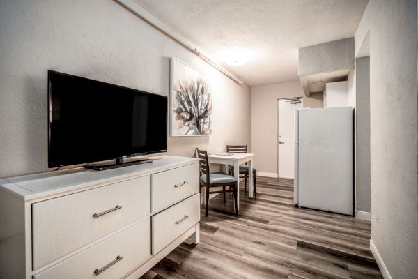 A small, neatly furnished room with a TV on a dresser, a dining table with chairs, artwork on the wall, and a refrigerator near the entrance.