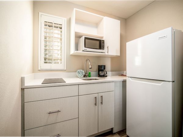 A compact kitchen with a fridge, microwave, sink, coffee maker, and cabinets, all in a clean, modern design with white and light wood tones.