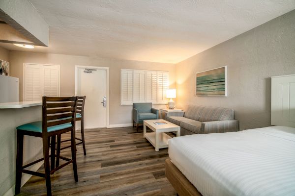 A modern hotel room with a bed, a sitting area, a table, a lamp, bar stools by a counter, and wall art, featuring wooden flooring and neutral tones.