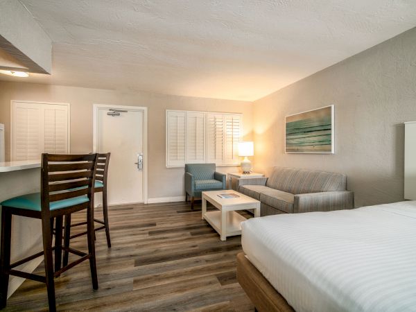 A modern hotel room with a bed, a sitting area, a table, a lamp, bar stools by a counter, and wall art, featuring wooden flooring and neutral tones.
