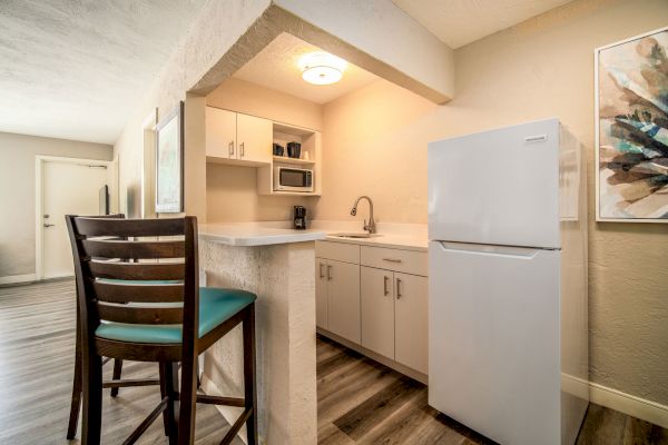 The image shows a small kitchen area with a fridge, sink, cabinets, and a small bar counter with two chairs in a modern apartment setting.