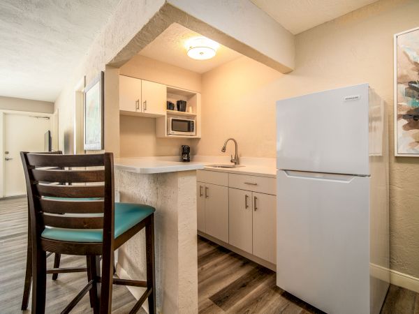 The image shows a small kitchen area with a fridge, sink, cabinets, and a small bar counter with two chairs in a modern apartment setting.