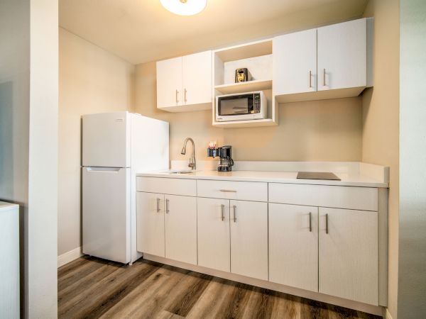 This image shows a compact modern kitchenette with white cabinets, a refrigerator, microwave, sink, and coffee maker on wooden flooring.