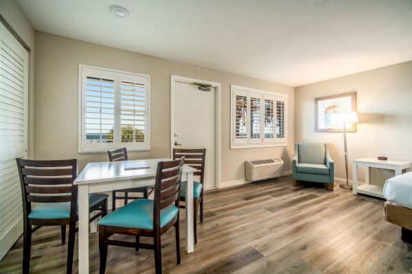 This image shows a cozy room with a dining table and four chairs, an armchair, a bed, and a lamp near a window with blinds.