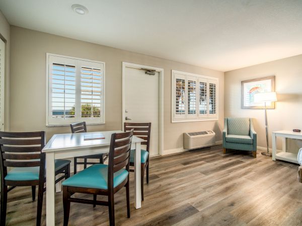 This image shows a cozy room with a dining table and four chairs, an armchair, a bed, and a lamp near a window with blinds.