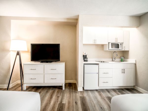 The image shows a hotel room with a flat-screen TV on a dresser, a kitchenette with white cabinets, a small fridge, a microwave, and a lamp.