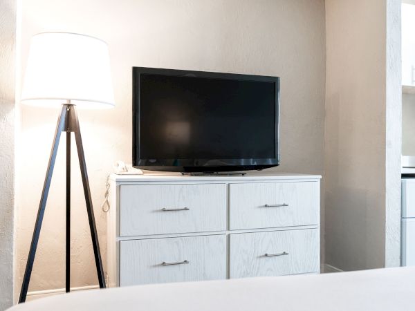 A black TV on a white dresser with four drawers next to a floor lamp with a white shade in a bright room.