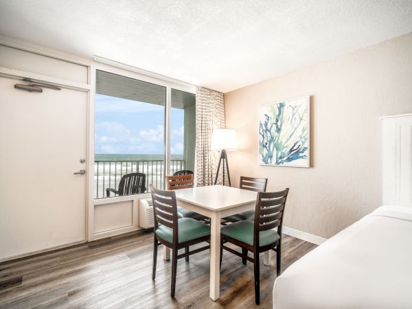 The image shows a bright room with a table and four chairs, a bed, a floor lamp, wall art, and a door leading to a balcony with an ocean view.