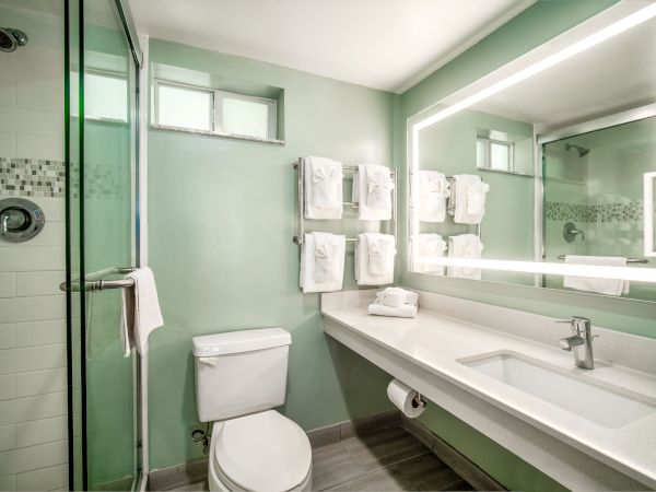A modern bathroom with a glass shower, toilet, and large illuminated mirror above a sink counter; towels are neatly hung on a rack.