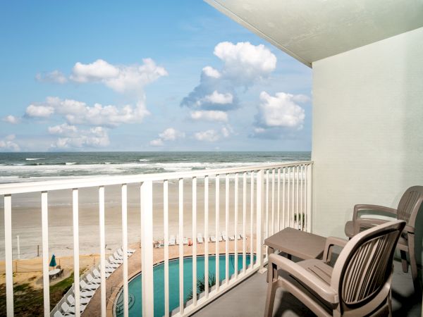 A balcony overlooking a beach, featuring a pool, two chairs, a small table, and the ocean under a blue sky with scattered clouds.