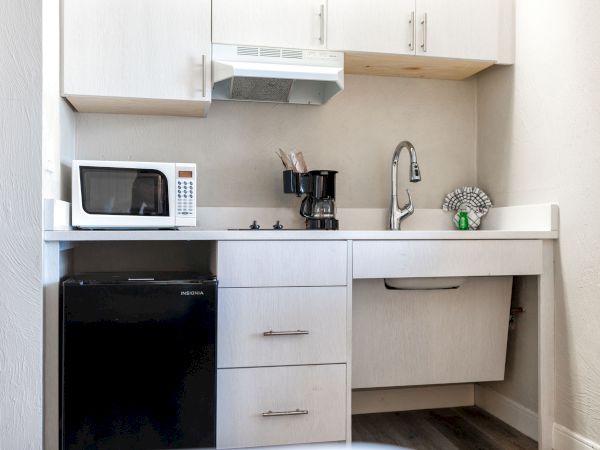 A compact kitchenette with a microwave, coffee maker, sink, faucet, small fridge, and cabinets above, all in a light-colored finish.