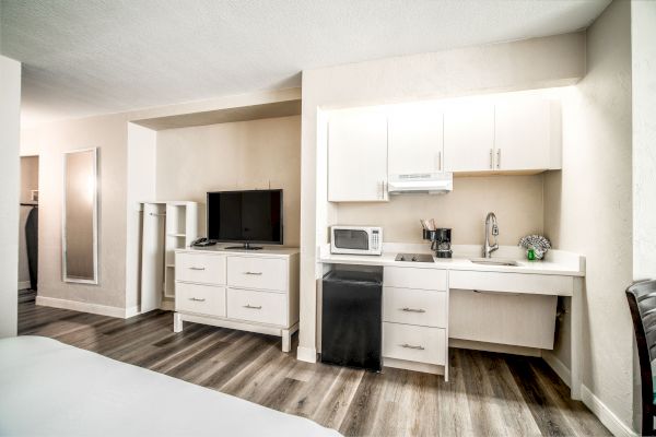 The image shows a small kitchenette with cabinets, a microwave, a coffee maker, a sink, a TV, and a dresser in a modern room.