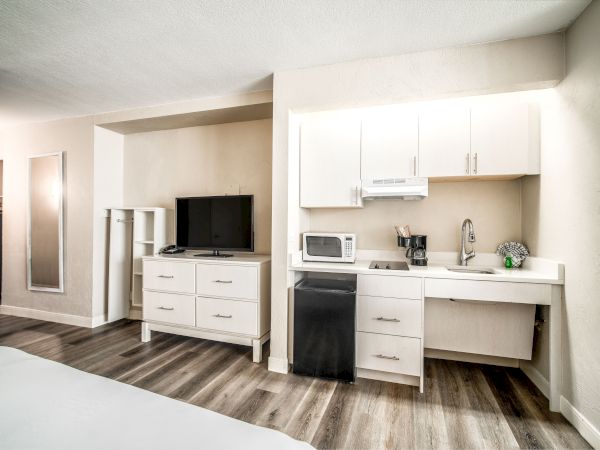 The image shows a small kitchenette with cabinets, a microwave, a coffee maker, a sink, a TV, and a dresser in a modern room.