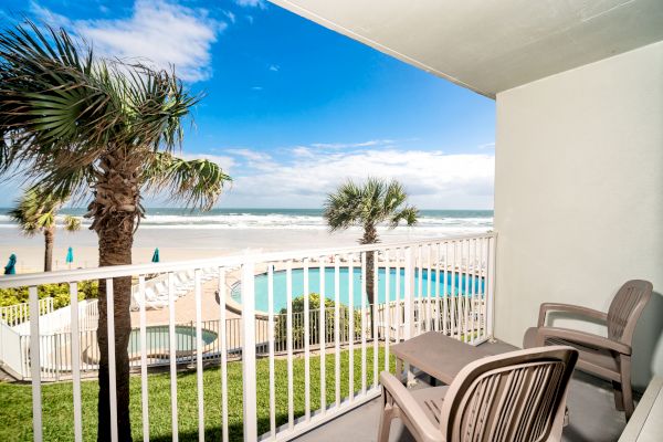 A balcony with two chairs overlooks a pool, palm trees, and a beach with the ocean in the background under a partly cloudy sky.