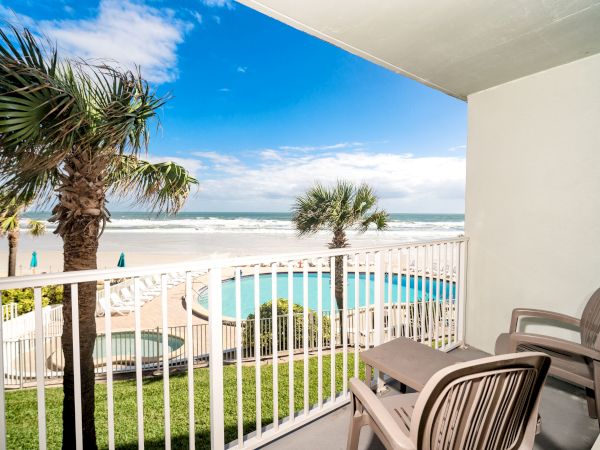 A balcony with two chairs overlooks a pool, palm trees, and a beach with the ocean in the background under a partly cloudy sky.