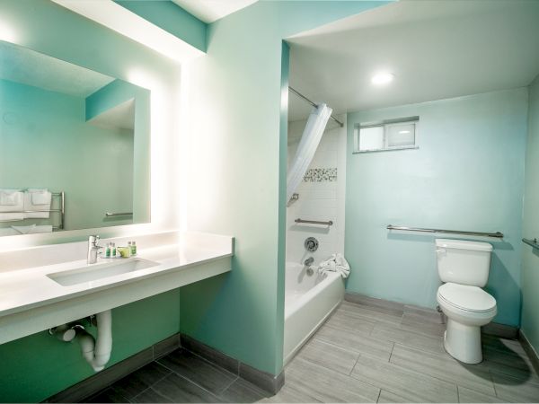 A modern bathroom with a large mirror, sink, toilet, bathtub with shower curtain, and teal walls. The room is well-lit with a minimalist design.