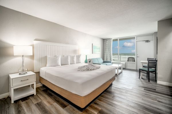 A modern hotel room with a king-sized bed, nightstands, a couch, a table, a balcony view of the ocean, and a swan towel on the bed.