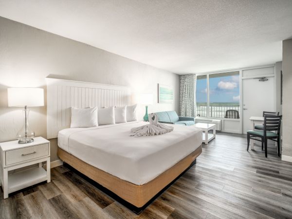 A modern hotel room with a king-sized bed, nightstands, a couch, a table, a balcony view of the ocean, and a swan towel on the bed.