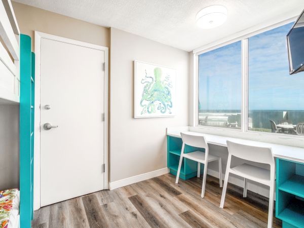 The image shows a bright room with a door, artwork of an octopus, windows with an ocean view, a desk with two chairs, and a wall-mounted TV.